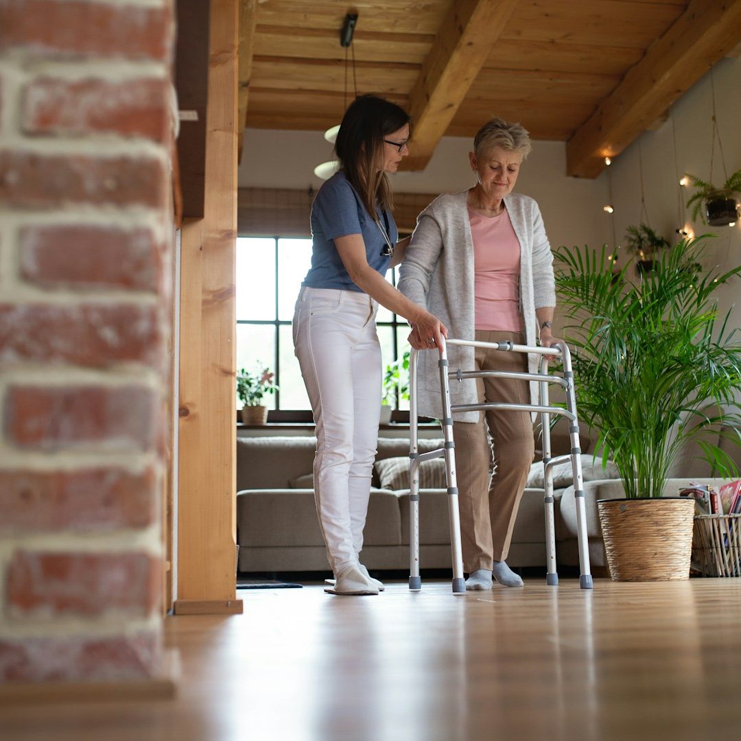 Eine aufmerksame Betreuungskraft begleitet eine Seniorin im häuslichen Umfeld sicher bei den ersten Schritten mit einer Gehhilfe.